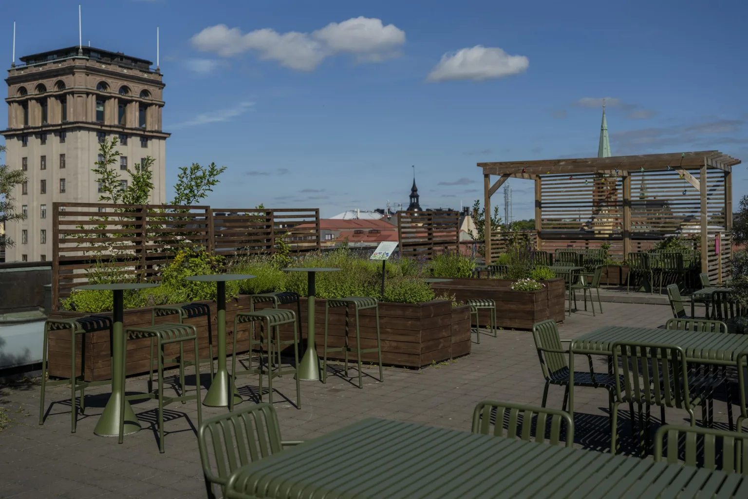 En takterrass med bord, stolar, växter i lådor och utsikt över stadens byggnader under en blå himmel.