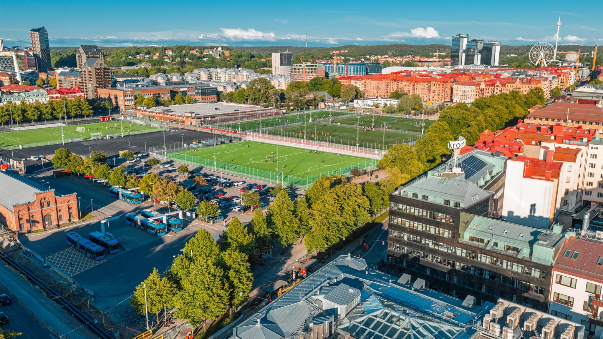 Flygbild av ett grönområde med fotbollsplaner omgivna av byggnader och träd, med en stad i bakgrunden.