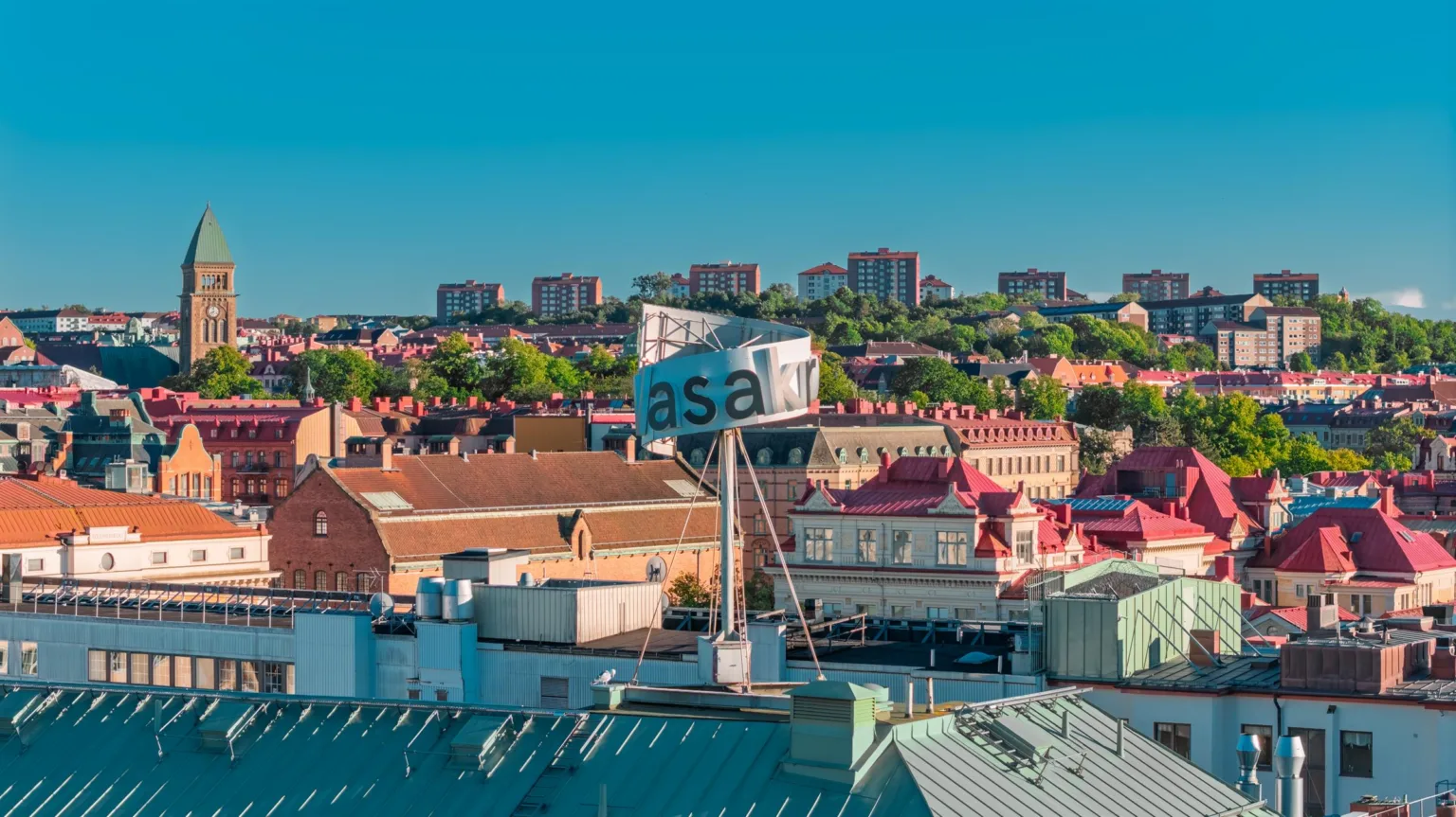 Skylt med texten "asa" på ett tak, omgiven av byggnader och grönska i bakgrunden.