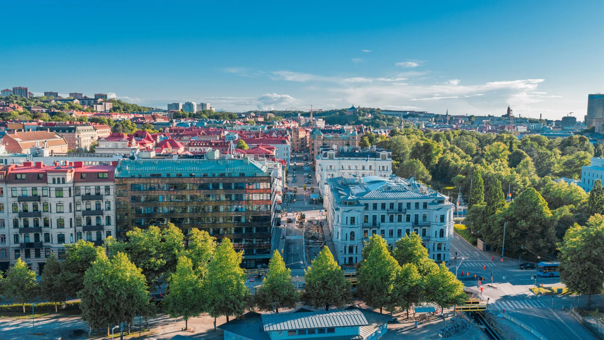 En stadsvy med färgglada byggnader, grönskande träd och blå himmel under en solig dag.