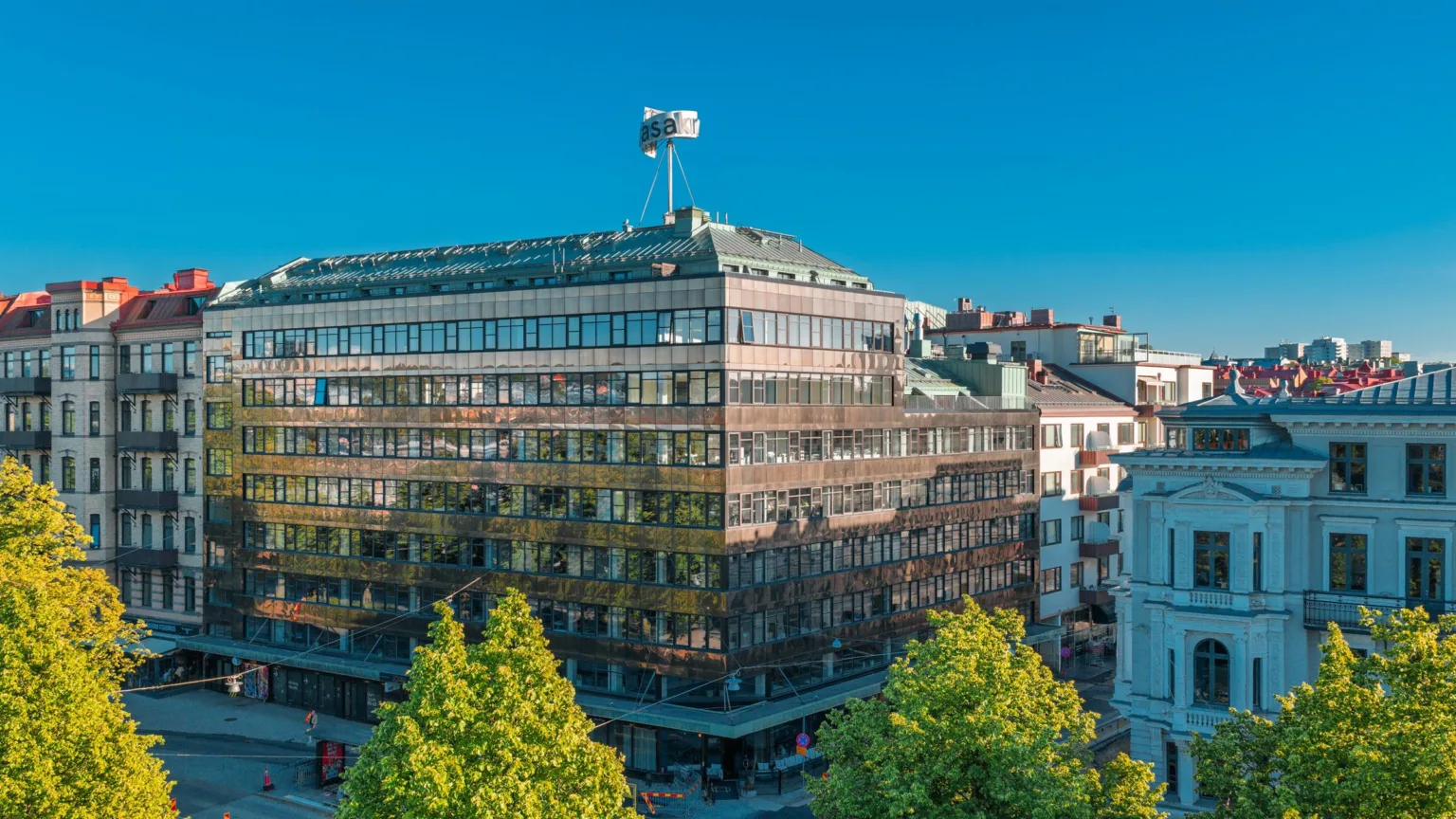 - En stor byggnad med glasfasad, omgiven av träd, under en klarblå himmel.