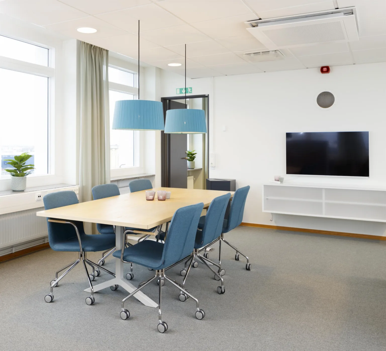 Konferensrum med bord, blå stolar, hängande lampor och en TV på en vit hylla.