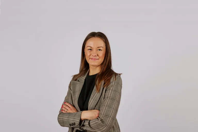 - En kvinna med långt brunt hår och en grå rutmönstrad kavaj korsar armarna och ler mot en ljus bakgrund.