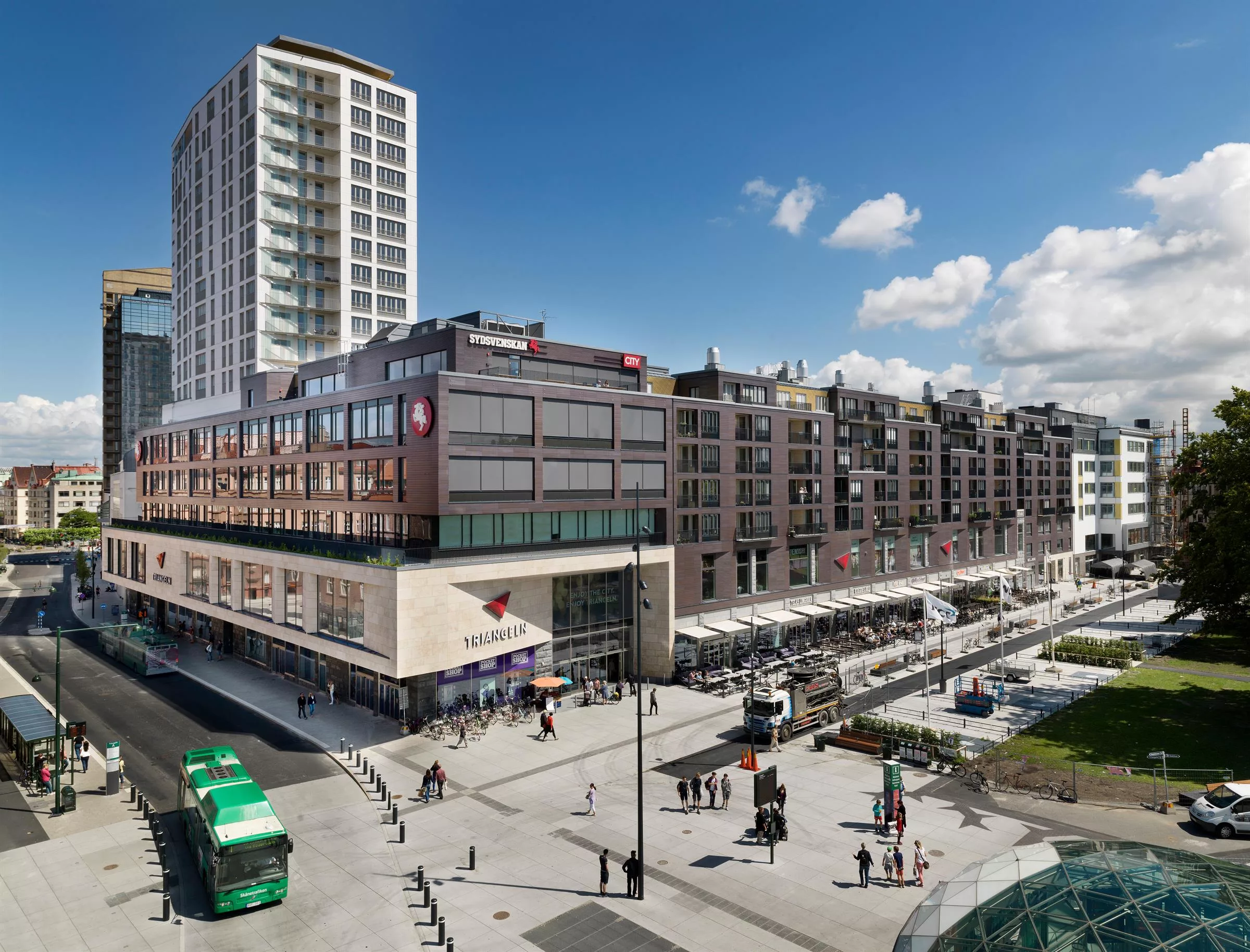 1. En stadsbild med en modern byggnad, höghus och gågata med människor och bussar under en klar blå himmel.