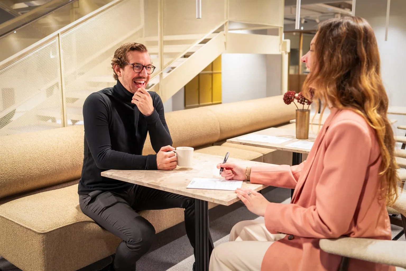 Två personer sitter vid ett bord och samtalar; en person håller en penna och antecknar medan den andra ler och håller en mugg.