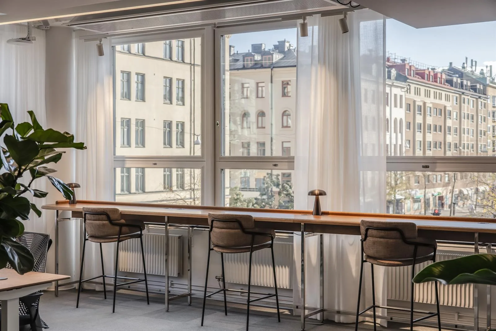 Ett långbord med höga stolar står framför stora fönster med utsikt över stadens byggnader.