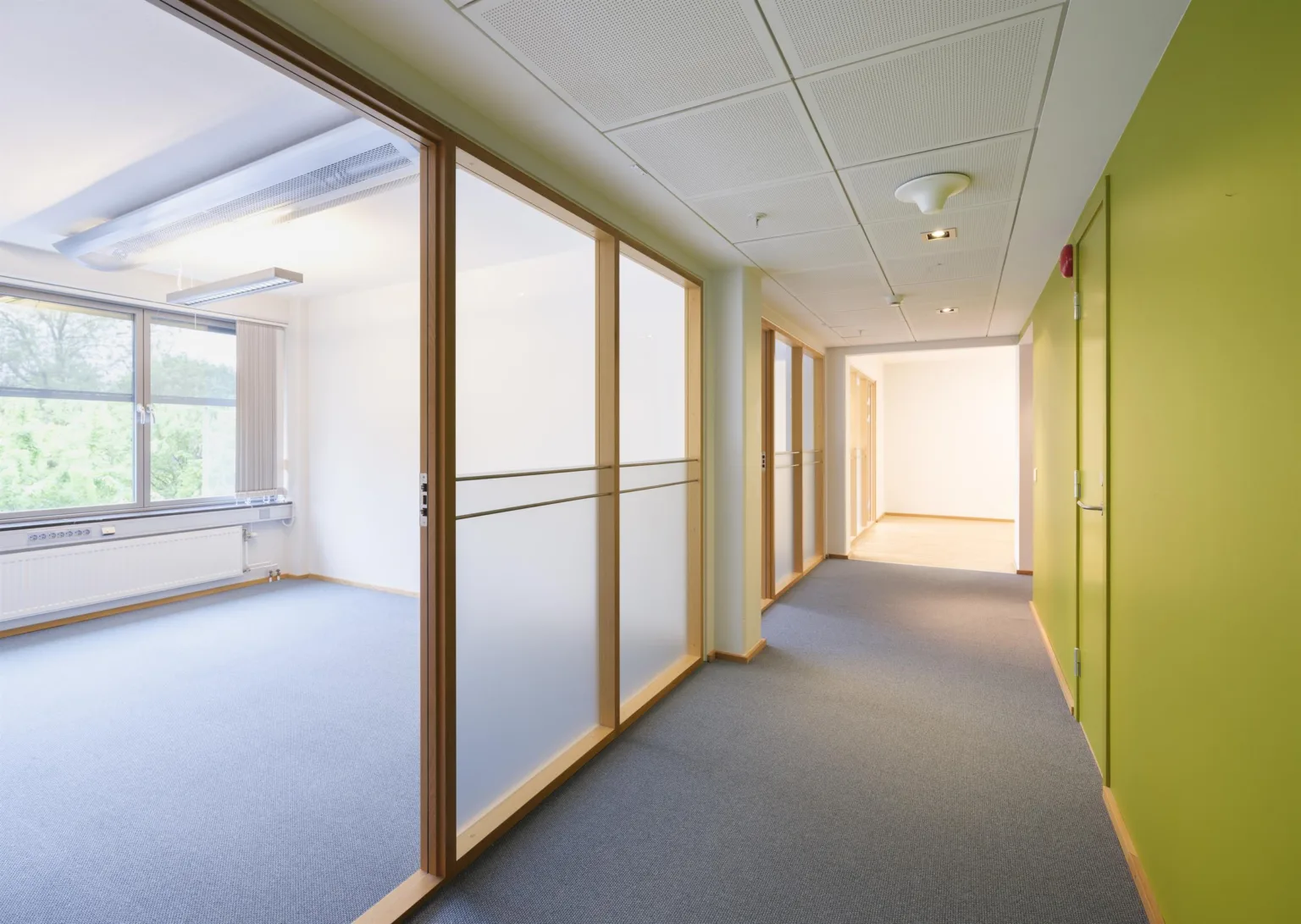 En annan korridor med glasväggar, träramar och gröna inslag på väggarna, som leder till ett upplyst rum.