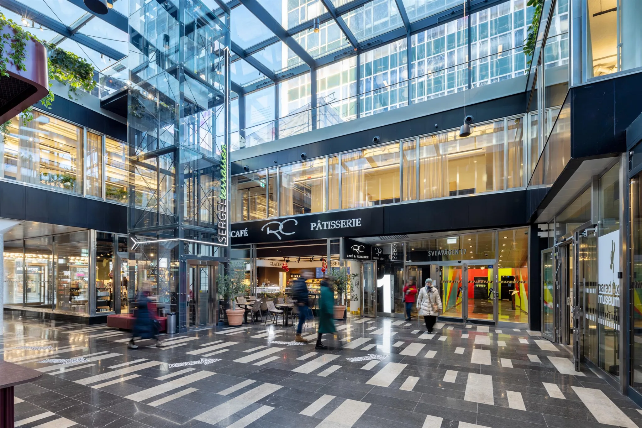 En ljus och rymlig galleria med glasväggar, butiker och människor som promenerar.