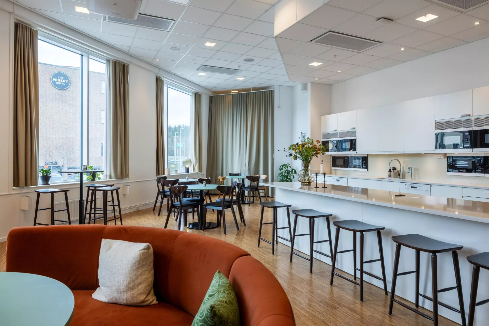 4. En lounge med en rund orange soffa, ett kök med vit köksö och pallar samt flera små bord och stolar vid fönstren.