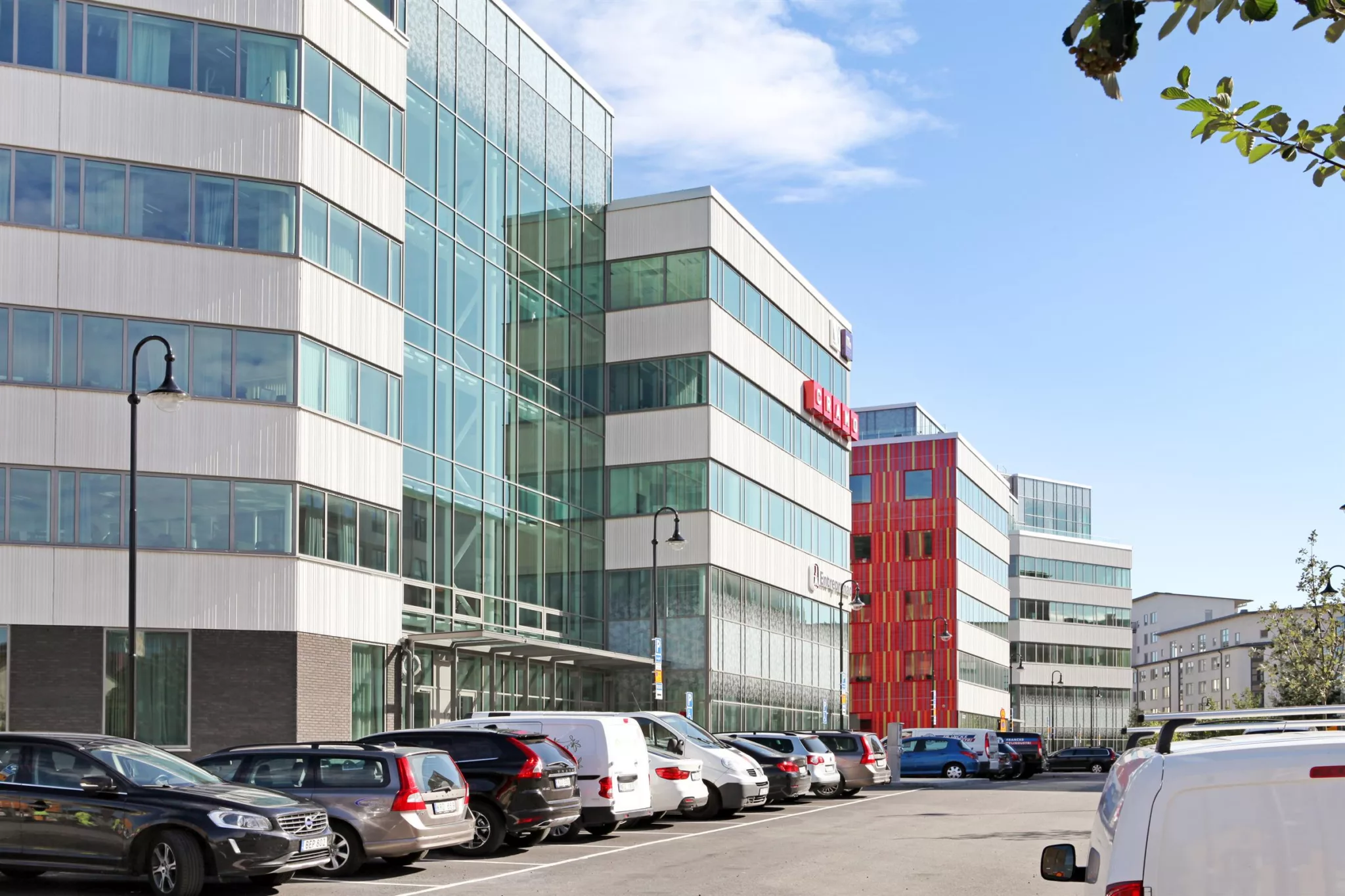 Gata med flera moderna kontorsbyggnader med glasfasader, parkerade bilar längs trottoaren och klarblå himmel.