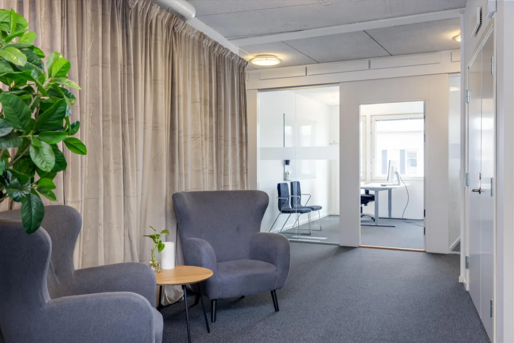 4. En lounge med två fåtöljer, ett litet bord och gardiner, med utsikt mot glasväggar och kontorsutrymmen.